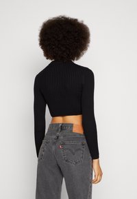 Top court noir côtelé à manches longues, porté avec un jean en denim gris taille haute, orné d'une écusson en cuir et d'un logo rouge à l'arrière.