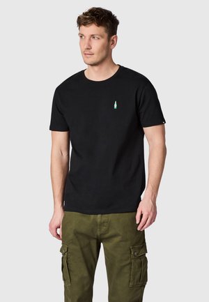 Man draagt een zwart T-shirt met een klein groen en wit fleslogo op de borst en olijfgroene cargobroek, staand tegen een lichte achtergrond.