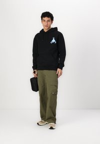 Black hoodie with kangaroo pocket and blue logo; olive cargo pants with pockets; beige sneakers; holding a small black bag.