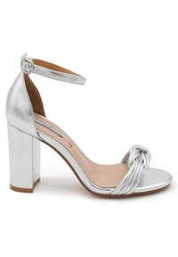 Sandaletto alto in metallo argentato con tacco a blocco quadrato, cinturino alla caviglia e dettaglio a nodo intrecciato nella zona delle dita. Texture liscia.