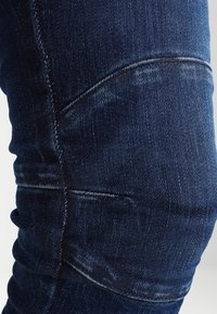 Mörkblå denimjeans med en figursydd design, som har synlig sömnad, subtila blekningar och sträv väv nära knäområdet.