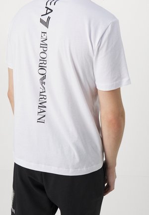 Tył osoby ubraną w białą koszulkę z krótkim rękawem z pionowym, czarnym logo „Emporio Armani”, w połączeniu z czarnymi spodniami.