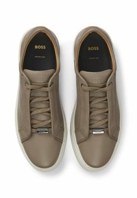 Un paio di sneaker in pelle taupe BOSS con superficie texture, lacci beige, suole bianche e logo dorato all'interno. Realizzate in Italia.