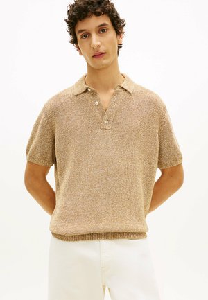 Giovane uomo con capelli ricci che indossa una polo beige a maniche corte con trama e pantaloni bianchi, in piedi con le mani dietro la schiena.