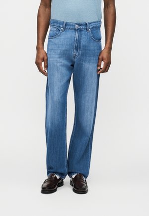 Mees, kes kannab helerohelisi sirge säärega teksapükse, helerohelist särke, mis on sisse volditud, valgeid sokke ja tumepruune nahast mokasiine.