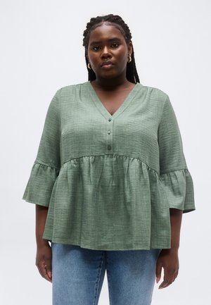 Γυναίκα με πλεξούδες, φορώντας ένα αέρινο ανοιχτό πράσινο πουκάμισο με μανίκια καμπάνα 3/4 και μπλε τζιν, στέκεται μπροστά σε ένα απλό λευκό φόντο.