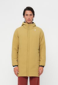 Gul parkas med hette, frontglidelås, sidelommer og elastiske mansjetter. Har et logo på brystet og et glatt, strukturert stoff.
