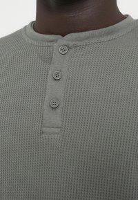 Grå vaffelsydd henley-skjorte med tre-knappersslå og ribbet krage, som fremhever det teksturerte stoffet og det avslappede designet.
