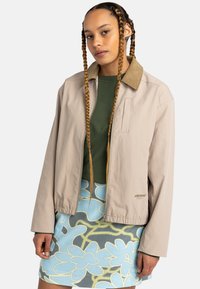 Lichtbeige jasje met een tan kraag, ritsluiting en een zak op de borst; gecombineerd met een groene blouse en een gebloemde blauwe rok.