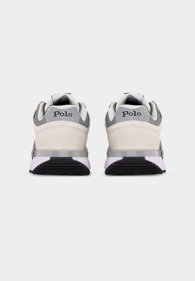 Vista trasera de unas zapatillas Polo grises y blancas que muestran un collar acolchado, el logo en la lengüeta del talón y suelas de goma negras sobre un fondo claro.