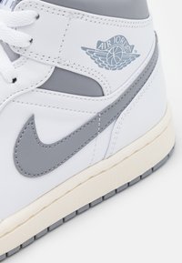 Gros plan sur une basket Air Jordan blanche et grise montrant les lacets, le swoosh Nike, le logo aile Air Jordan et la semelle crème.