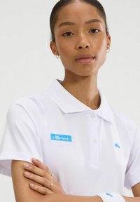 Camiseta polo blanca con un cuello clásico, que presenta un logotipo azul de "ellesse" en la parte superior izquierda y un pequeño acento azul cerca del cuello.