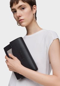 Black textured leather clutch with embossed "TOUS" logo, featuring a flat design and a contrasting light interior, held in a hand.