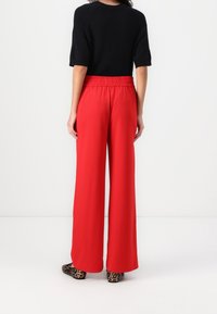 Pantalon rouge à jambes larges avec une ceinture élastique, fabriqué dans un tissu lisse. Porté avec un haut noir et des chaussures à motif léopard.