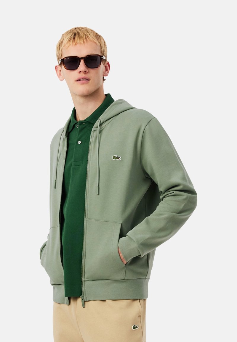 Sudadera con capucha verde con cremallera y bolsillo canguro, combinada con una camiseta polo verde oscuro. El modelo lleva gafas de sol negras y pantalones beige. Logo de Lacoste visible.