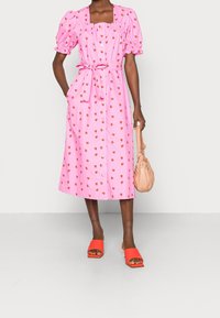 Robe rose, à mi-cuisse, avec un décolleté carré, des manches bouffantes et un motif de fraises. Associée à des chaussures orange à enfiler et un sac à main beige.