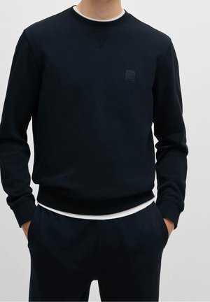 Man draagt een zwart langarmige trui en bijpassende zwarte broek met handen in de zakken tegen een effen lichte achtergrond.
