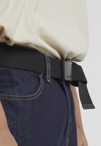 Črn najlon pas z pravokotno črno sponko, nošen čez temno modre jeans hlače z rumenim šivom. Teksturiran material, nastavljive dolžine.
