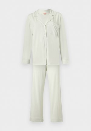 Femme debout pieds nus portant une chemise ample claire à manches longues boutonnée et un pantalon large assorti sur un fond uni.