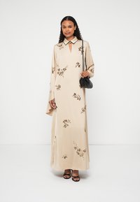 Beige virágmintás maxi ruha, kulcslyuk kivágással és fekete gallérral. Hosszú ujjakkal rendelkezik és sima, légies textúrája van. Kiegészíti egy fekete táska.