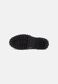 Semelle de chaussure en caoutchouc noir présentant une surface texturée avec de profonds cannelures et un détail de logo. Conçue pour l'adhérence avec une forme épaisse.