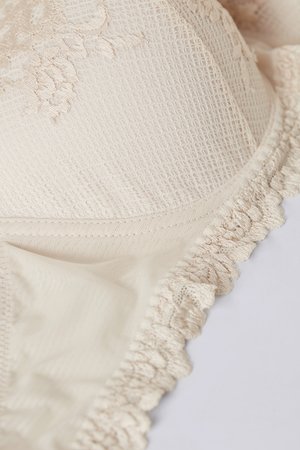 Béžová krajková podprsenka s texturovanou látkou, květinovou výšivkou a volánkovými okraji, která představuje hladký a jemný design.