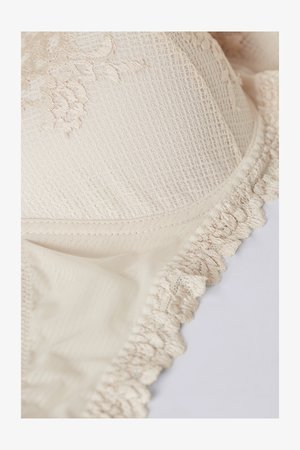 Béžová krajková podprsenka s texturovanou látkou, květinovou výšivkou a volánkovými okraji, která představuje hladký a jemný design.