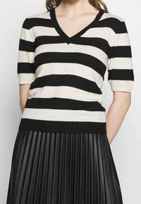 Žena v černobíle pruhovaném topu s V výstřihem a černé skládáním sukně, stojící s uvolněnými rukama za zády.