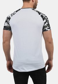 Weißes T-Shirt mit camouflagefarbigen Raglanärmeln in Grautönen und Schwarz. Hergestellt aus weichem Material, mit taillierter Passform und kurzen Ärmeln.