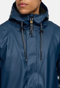 Navyblauwe regenjas met een structuurovervlak, verstelbare capuchon, zwart-witte trekkoorden en een ritsopening aan de voorkant.