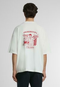 Fehér, oversized póló, hátul piros grafikával, amelyen egy pizzaiolo és egy pizzéria látható, valamint szöveggel és kockás mintával.