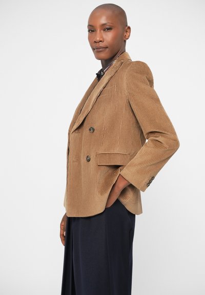 Rjav kordurojski dvoredni blazer z odrezanimi klapnicami, dvema sprednjima žepoma in gumbi. Kombiniran s temno modrimi hlačami.