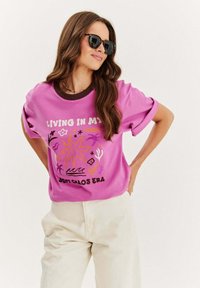 Růžové bavlněné tričko s hnědým výstřihem a grafickým designem obsahujícím text a abstraktní prvky v žluté, fialové a černé barvě.