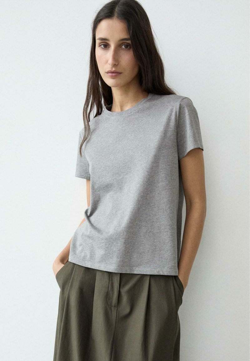 Vrouw met lang donker haar, gekleed in een effen grijs T-shirt met korte mouwen en olijfgroene broek, met handen in haar zakken, staand tegen een witte muur.