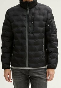 Černá prošívaná péřová bunda s vysokým límcem a několika kapsami na zip. Má šedé detaily a kapsu na rukávu s logem.