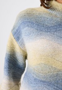 Close-up van een gebreide trui met getextureerde golfpatronen in blauw, beige en crèmekleuren op de bovenste torso van een persoon.