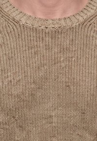 Gros plan d’un pull tricoté beige avec col côtelé visible et motif de points texturé.
