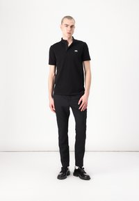 Juodas polo marškinėliai su trumpu apykaklės kraštu, tiesaus kirpimo, ir maža logotipas ant kairiojo krūtinės, derinami su siaurais juodais kelnėmis ir juodais batais.