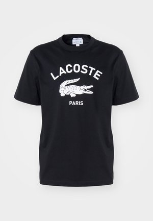 T-shirt noir à manches courtes avec un logo blanc Lacoste, représentant un crocodile à la bouche ouverte et le mot Paris en dessous.