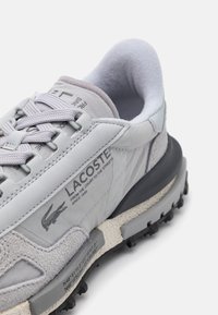 Light gray sneaker featuring a mix of textured materials, round laces, and a cushioning sole with black rubber tread and branding details.