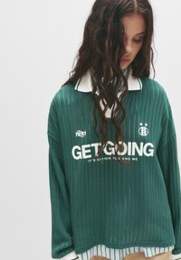 Zelená rebrová mikina s bielym nápisom "GET GOING" a golierom. Nosená na košeľu s golierom so zelenými a bielymi pruhmi na leme.