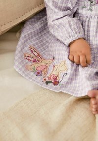 La mano de un niño descansa sobre un vestido de cuadros lilas decorado con conejitos bordados y detalles florales, sentado sobre una tela beige texturizada.