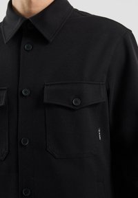 Czarna koszula na guziki wykonana z teksturowanej tkaniny. Posiada klasyczny kołnierzyk, dwie kieszenie na piersi oraz czarne guziki z subtelnym logo po boku.