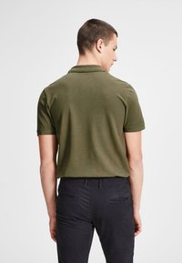 Olivgrünes Poloshirt mit Kragen, kurzen Ärmeln und glatter Textur, getragen mit dunklen Hosen. Von hinten betrachtet.