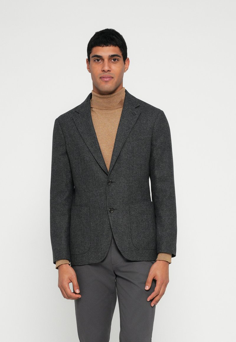 Blazer cinzento de mistura de lã com lapela entalhada, dois bolsos frontais e dois botões. Usado por cima de uma camisola de gola alta castanha, combinado com calças cinzentas.