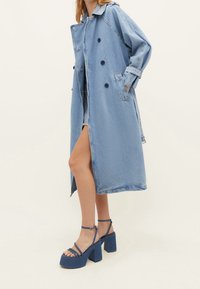 Ljusblå denim trenchcoat med dubbla knappar, stora knappar och sidofickor; ihopparad med kraftiga blå plattformssandal.