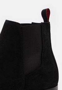 Bottine en daim noir avec panneaux latéraux élastiques, dotée d'une languette de tirage noir et rouge. Texture lisse et design élégant mettent en valeur les détails de la construction.