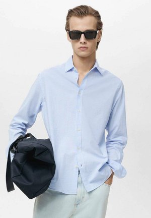 Light blue striped button-up shirt with long sleeves, worn with a casual light-colored bottom, holding a black bag and wearing dark sunglasses.