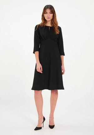 Robe noire avec des manches trois-quarts, coutures cintrées au niveau du corsage et jupe arrivant au genou. Fabriquée en tissu doux avec un léger éclat.