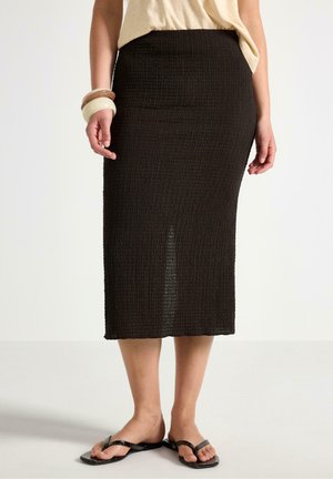 Kvinde iført en struktureret, sort midi-nederdel, beige top, to armbånd på venstre håndled og sorte sandaler med firkantet tå mod en ensfarvet baggrund.